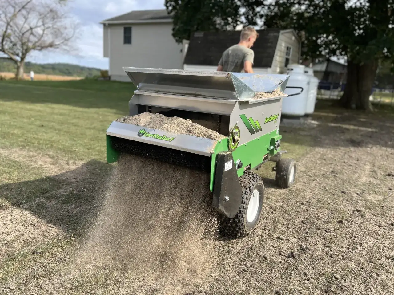 Ein Mann nutzt einen Streuwagen, um Rasensand auf einer Wiese zu verteilen. Was ist Rasensand? Dieses Material verbessert die Bodenstruktur und Drainage.