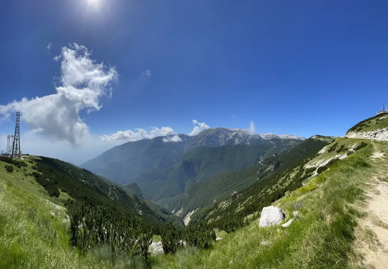 Panoramica mozzafiato sui monti dell'Abruzzo, con un sentiero che sale verso un **block house** e torri di trasmissione.