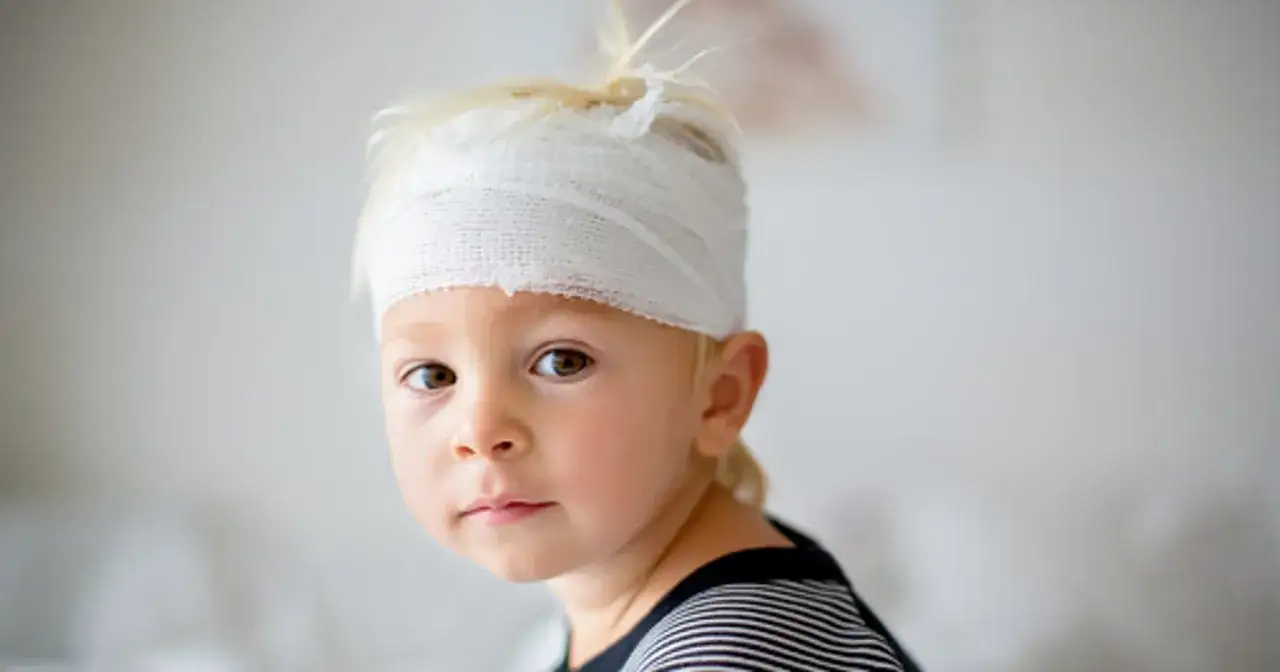 Mały chłopiec z blond włosami ma na głowie bandaż, co może sugerować wstrząs mózgu u dziecka.