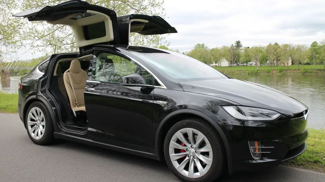 Czarny Tesla X z otwartymi drzwiami typu falcon wing i jasnymi skórzanymi siedzeniami, zaparkowany nad rzeką.