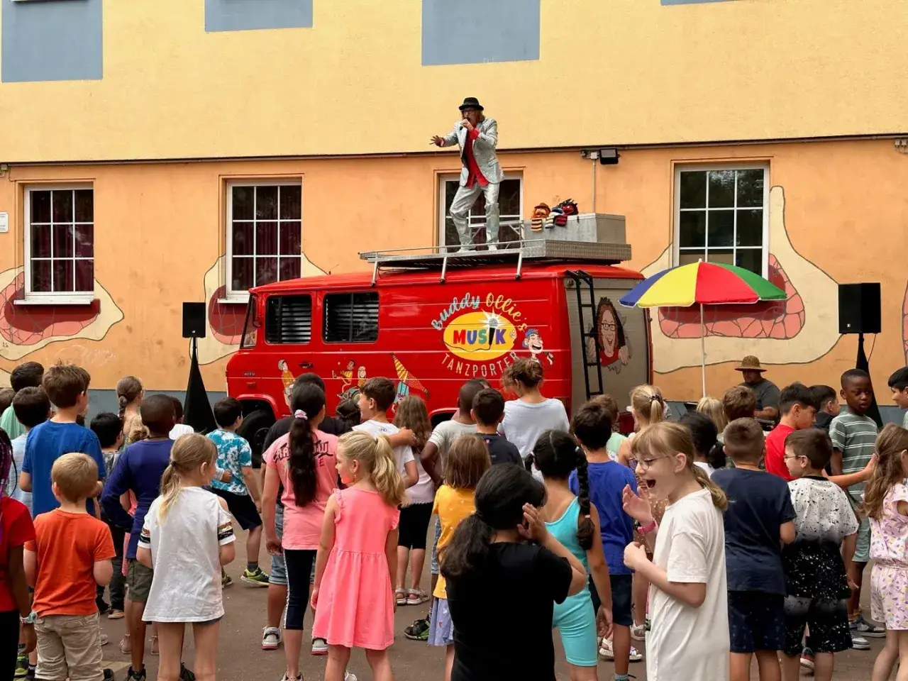 Ein Mann in einem glitzernden Anzug singt auf einem roten Musik-LKW. Kinder jubeln ihm zu. Ein fr&ouml;hlicher letzter Schultag Grundschule!