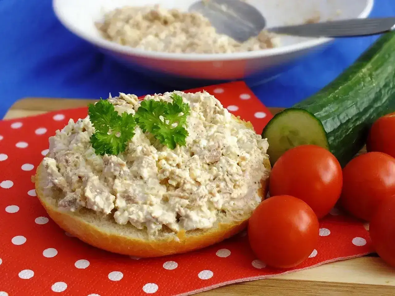 Pyszna pasta z tuńczyka i twarogu na bułce, udekorowana natką pietruszki. Obok pomidorki i ogórek.