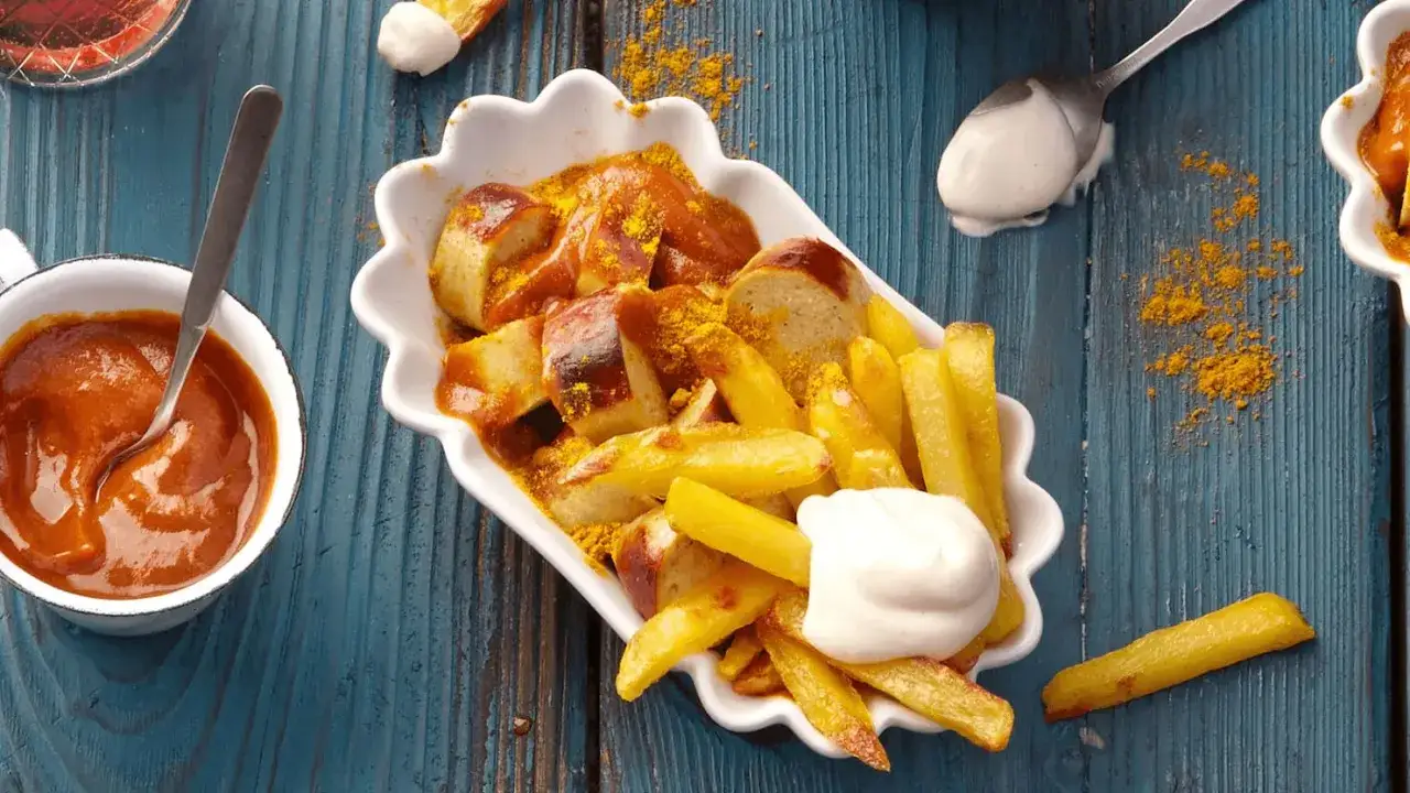 Eine Portion Currywurst mit Pommes und Mayonnaise. Die Currywurst ist in Scheiben geschnitten und mit Currypulver bestreut, die Pommes sind goldbraun.