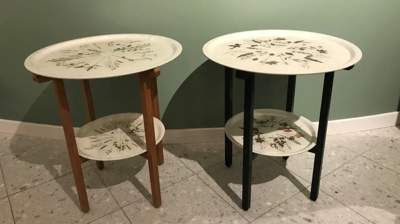 Två brickbord med trädetaljer och blomstermönster. Ett har en ljus trästomme, det andra en svart.