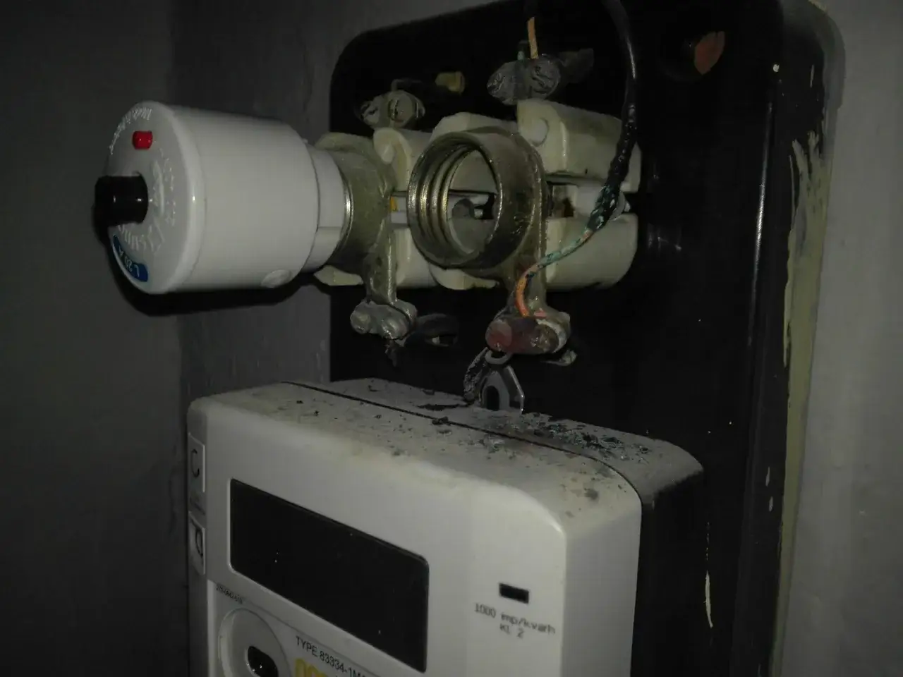 Widoczny przepalony bezpiecznik topikowy z czerwonym wskaźnikiem, obok licznik energii elektrycznej.