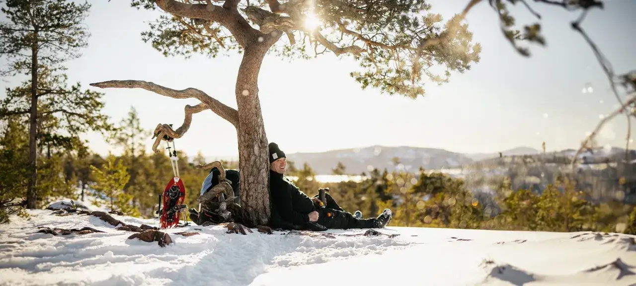 Man vilar under ett tr&auml;d i sn&ouml;n, redo f&ouml;r att vandra p&aring; vintern. Sn&ouml;skor och ryggs&auml;ck st&aring;r bredvid.