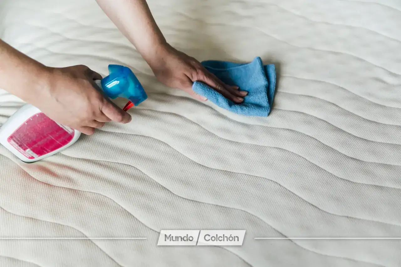 Manos rociando desinfectante y limpiando un colchón con un paño azul. Aprende como desinfectar un colchon para un descanso saludable.