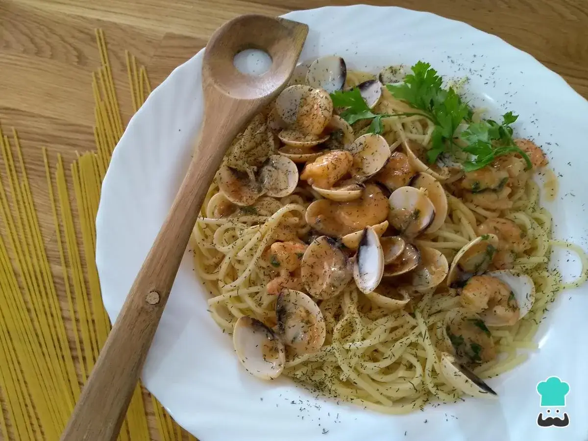 Espaguetis con gambas y almejas, servidos en un plato blanco con perejil y eneldo. Una cuchara de madera reposa a un lado.