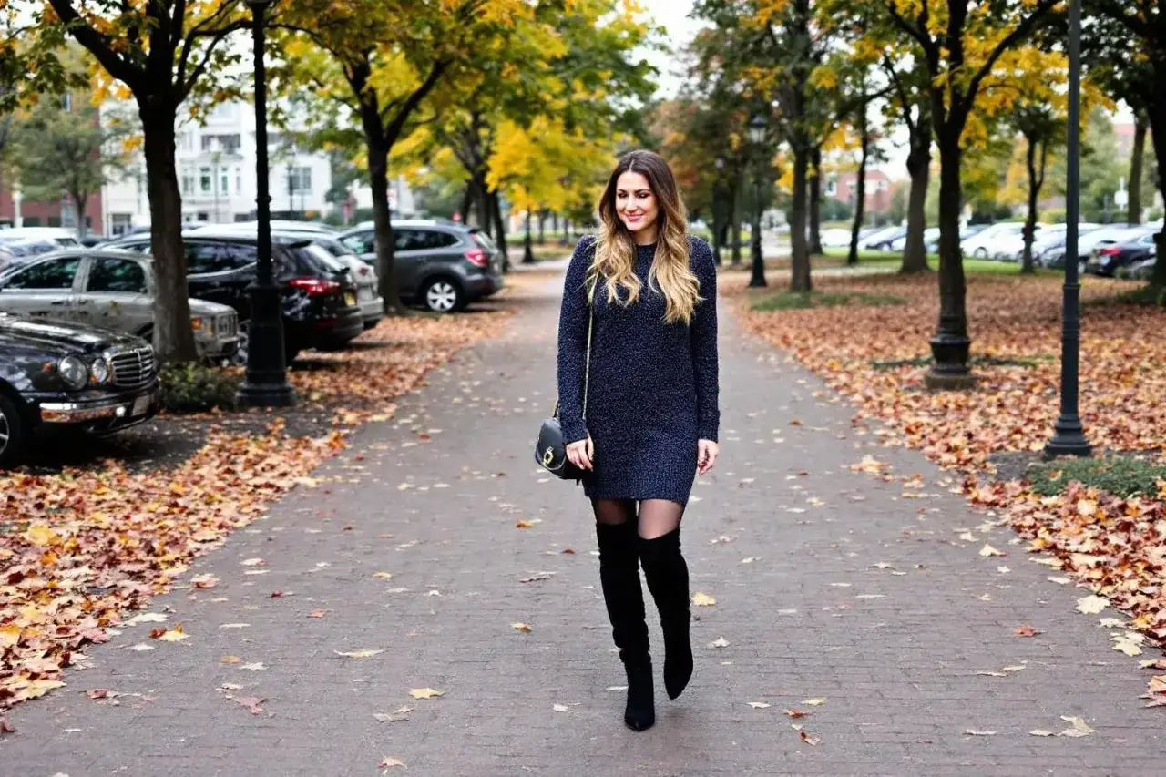 Une femme en robe pull bleu marine et cuissardes noires, parfaite pour savoir quelle chaussure mettre avec une robe pull en automne.