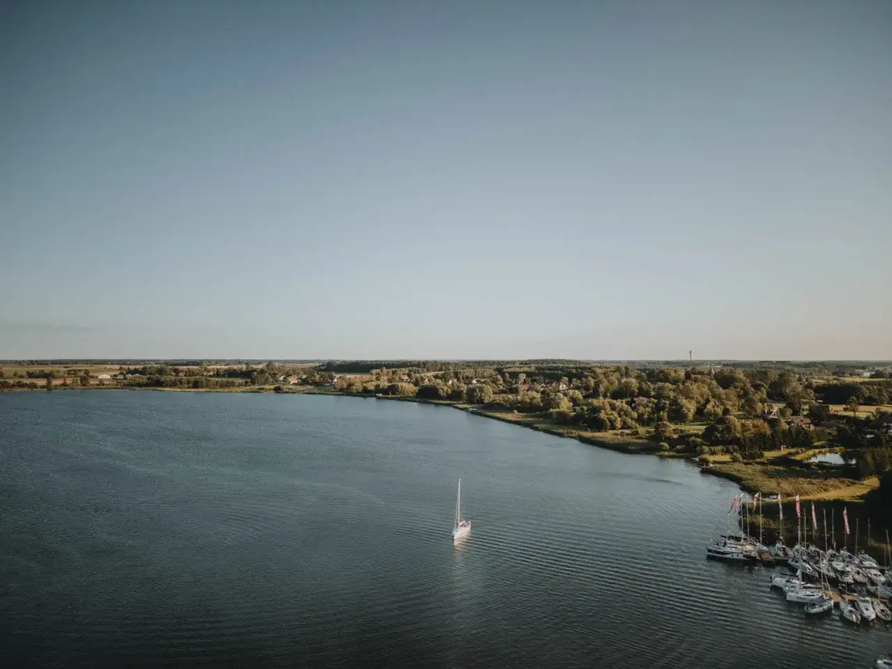 Spokojne jezioro Szymoneckie z samotnym żaglem na wodzie i malowniczą mariną pełną łodzi.