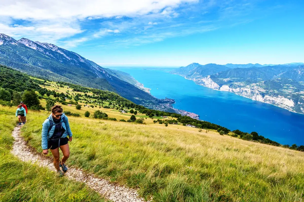Zwei Wanderer genießen die Aussicht beim Monte Baldo Wandern. Der Weg führt über einen sonnigen Hang mit Blick auf den tiefblauen Gardasee.
