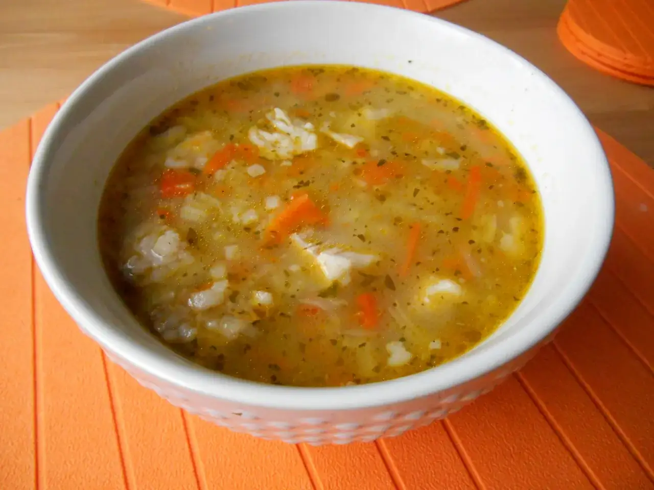 Aromaticzna zupa na żeberkach z ryżem i marchewką, podana w białej misce na pomarańczowej podkładce.