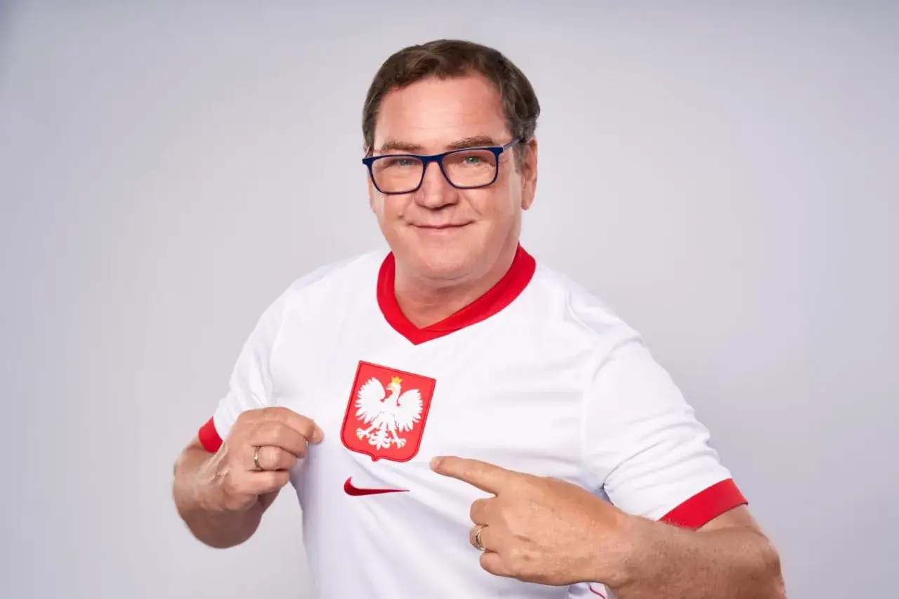 Uśmiechnięty mężczyzna w okularach wskazuje na herb Polski na swojej koszulce, jakby był trenerem reprezentacji Polski.