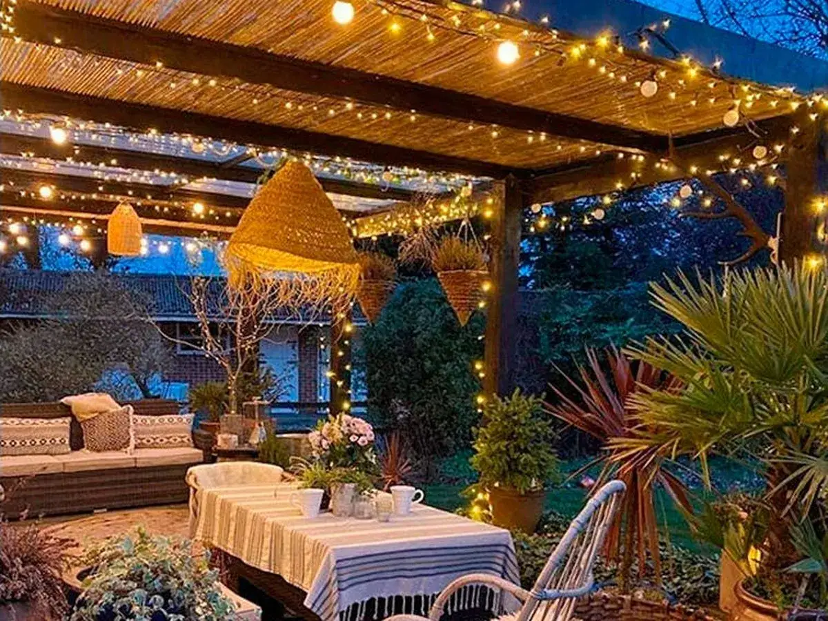 Un porche acogedor con luces c&aacute;lidas, ideal para saber como iluminar un jardin. Muebles de exterior, plantas y una mesa puesta crean un ambiente m&aacute;gico.