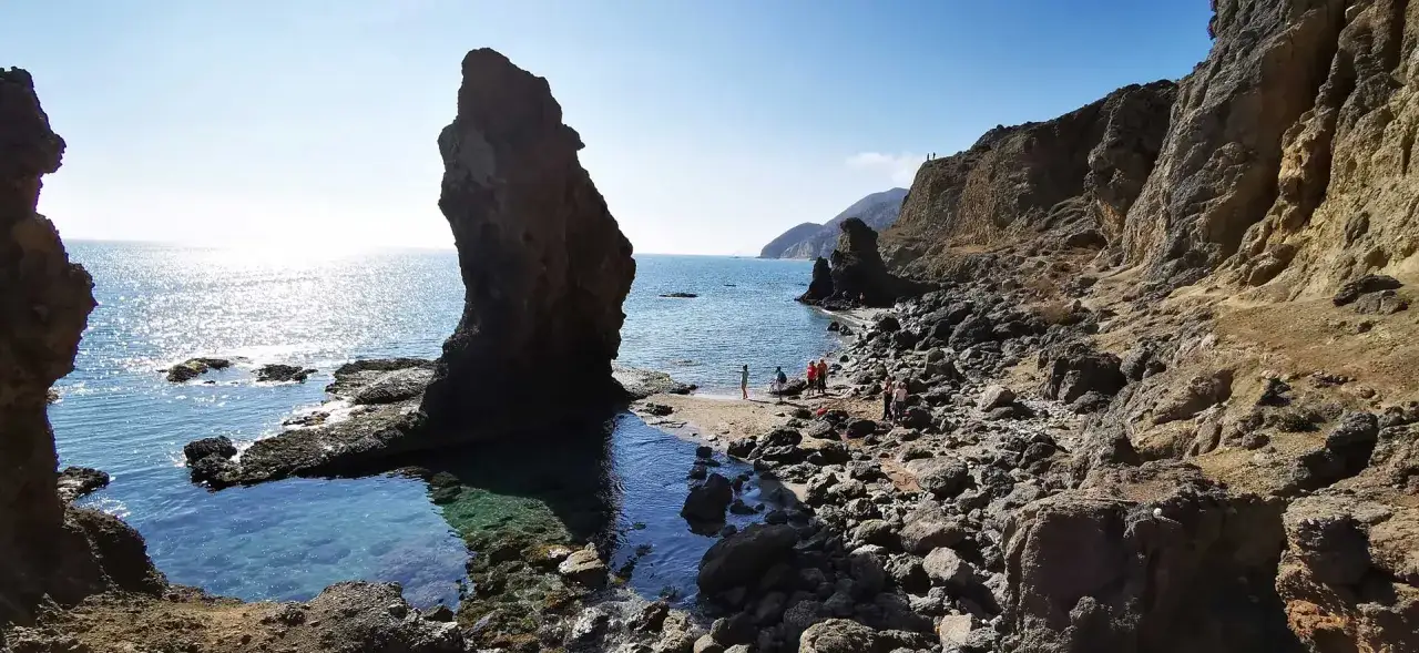 Gente disfruta de la **playa del Sombrerico**, con su imponente roca y aguas cristalinas. ¡Un lugar para guardar en la memoria!