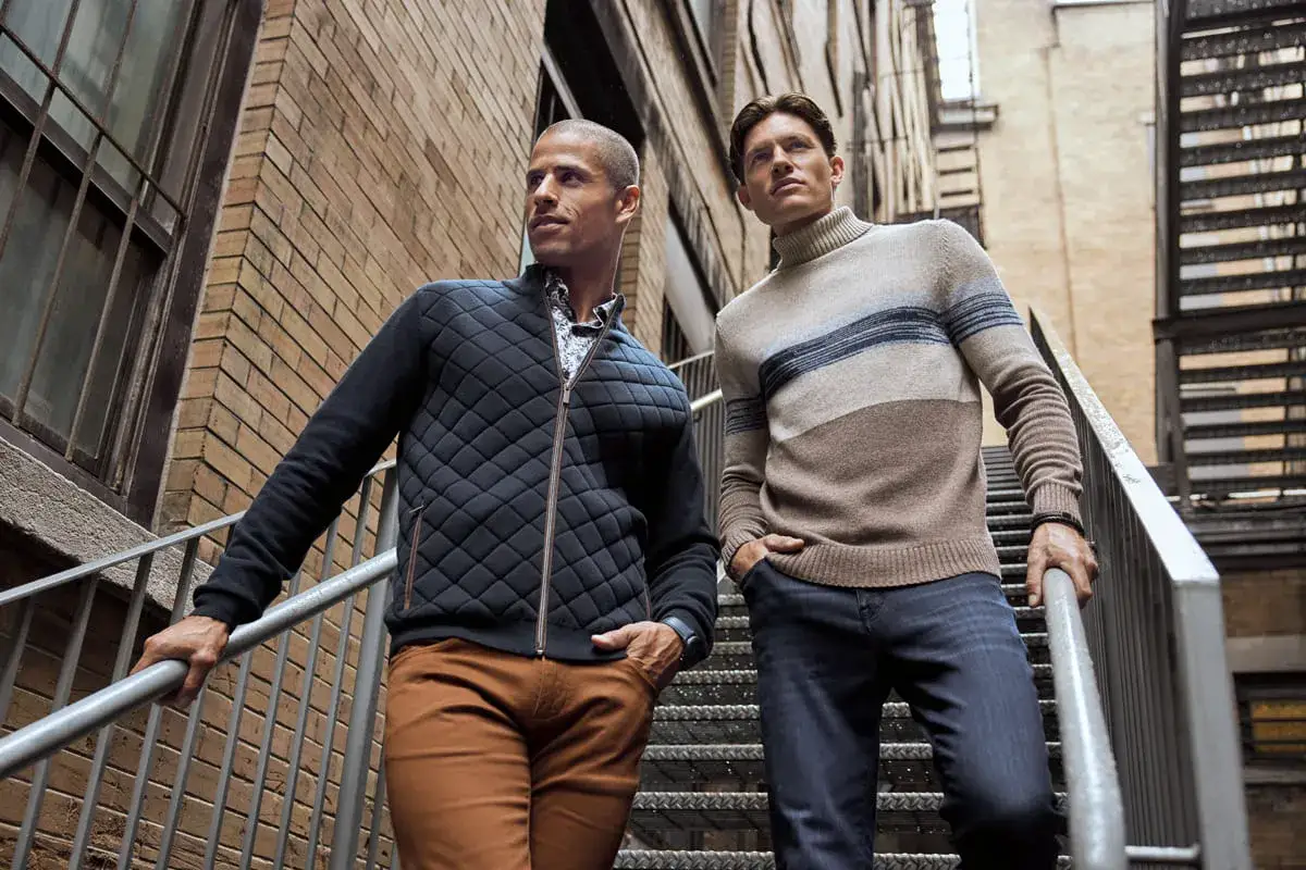 Zwei M&auml;nner auf einer Treppe, die den **casual chic dress code** verk&ouml;rpern. Einer tr&auml;gt eine gesteppte Jacke, der andere einen gestreiften Rollkragenpullover.