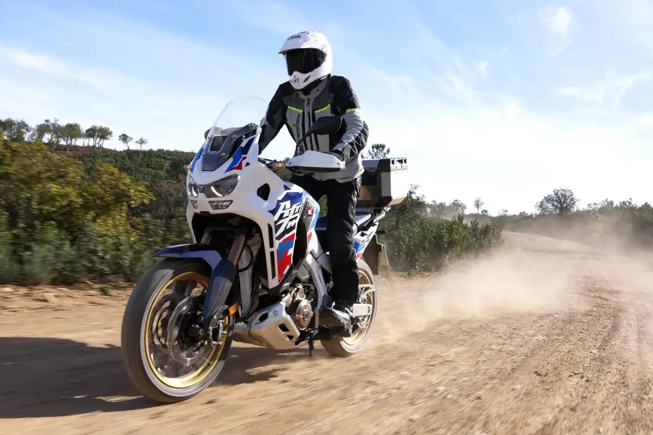 Motocyklista na Honda Africa Twin 1100 z kufrem pokonuje piaszczystą drogę, wzbijając tumany kurzu.