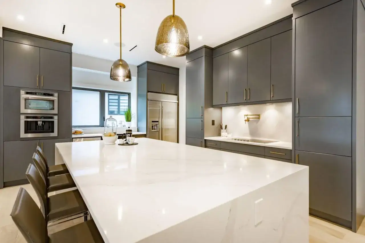 Cocina moderna con isla de cuarzo blanco. Dos l&aacute;mparas colgantes doradas iluminan el espacio, mostrando c&oacute;mo iluminar una cocina con estilo.