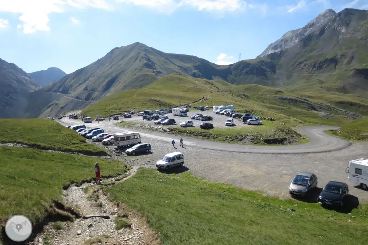 Un aparcamiento en la **col de Tentes** con coches y caravanas, rodeado de montañas verdes y un cielo azul.