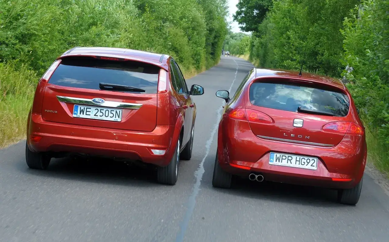 Dwa czerwone samochody, Ford Focus i Seat Leon, jadą obok siebie po wiejskiej drodze. Manewr wymijania w pełnym słońcu.