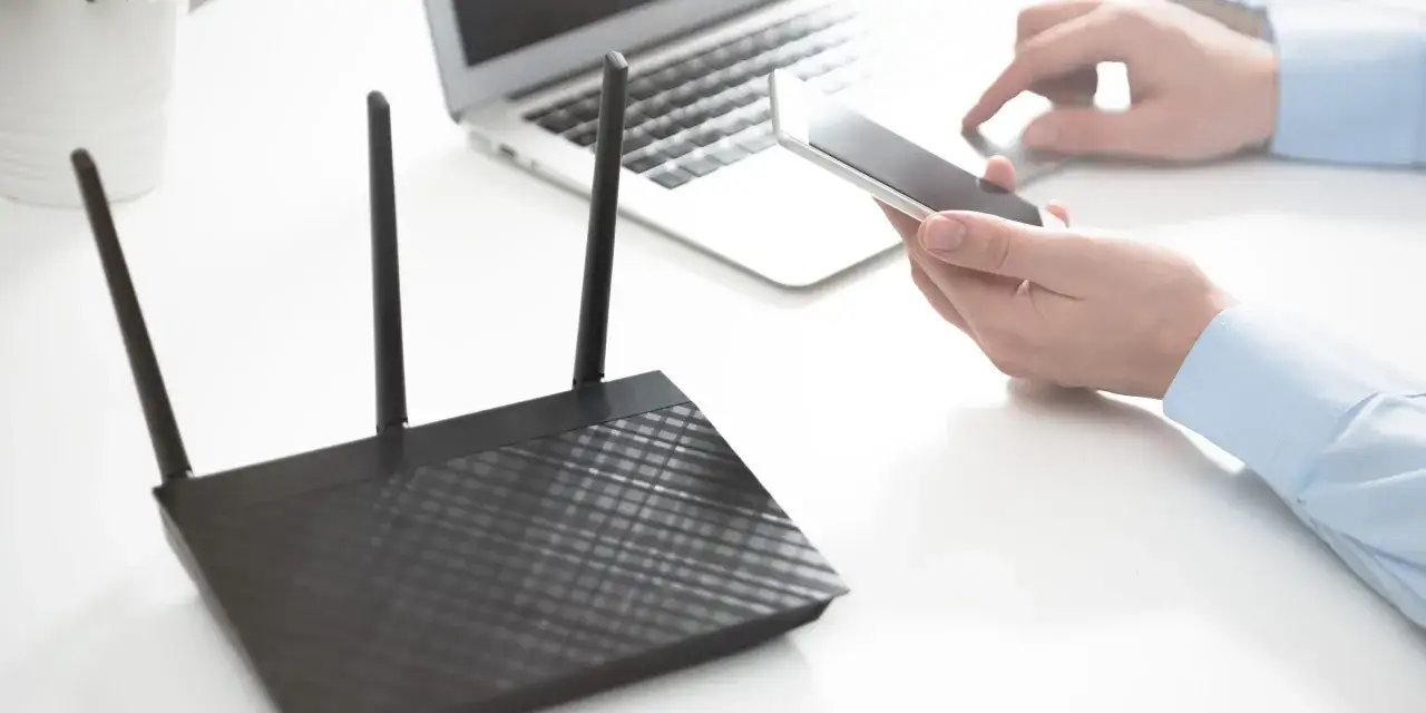 Czarny ruter z antenkami stoi obok laptopa. Dłoń trzyma smartfon, gotowa do połączenia z siecią.