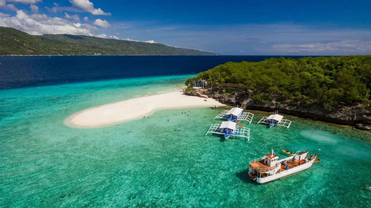 Vacker sandbank och turkost vatten vid Cebu, Filippinerna. B&aring;tar guppar i det klara vattnet, en gr&ouml;nskande &ouml; i bakgrunden.