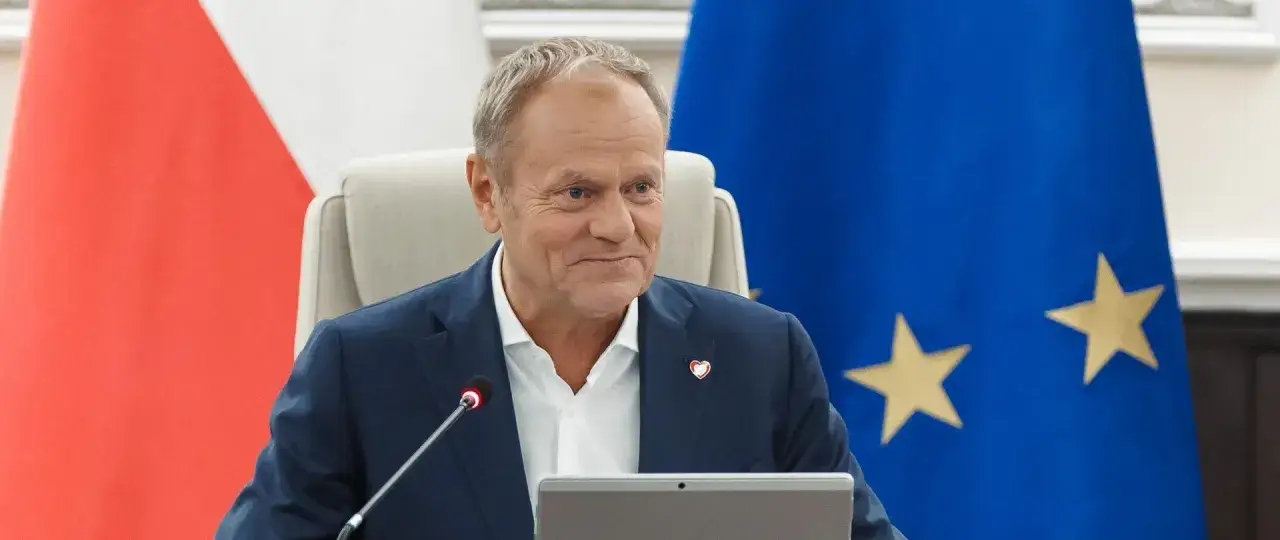 Uśmiechnięty Donald Tusk siedzi przed flagami Polski i UE. Czy to zapowiedź, kiedy kończy się kadencja premiera Tuska?