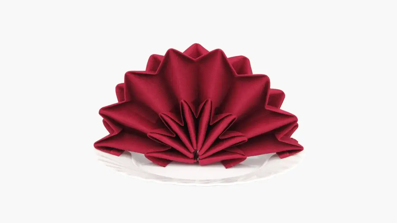 Une serviette rouge est pliée en éventail sur une assiette blanche. Ce pliage serviette éventail ajoute une touche d'élégance.
