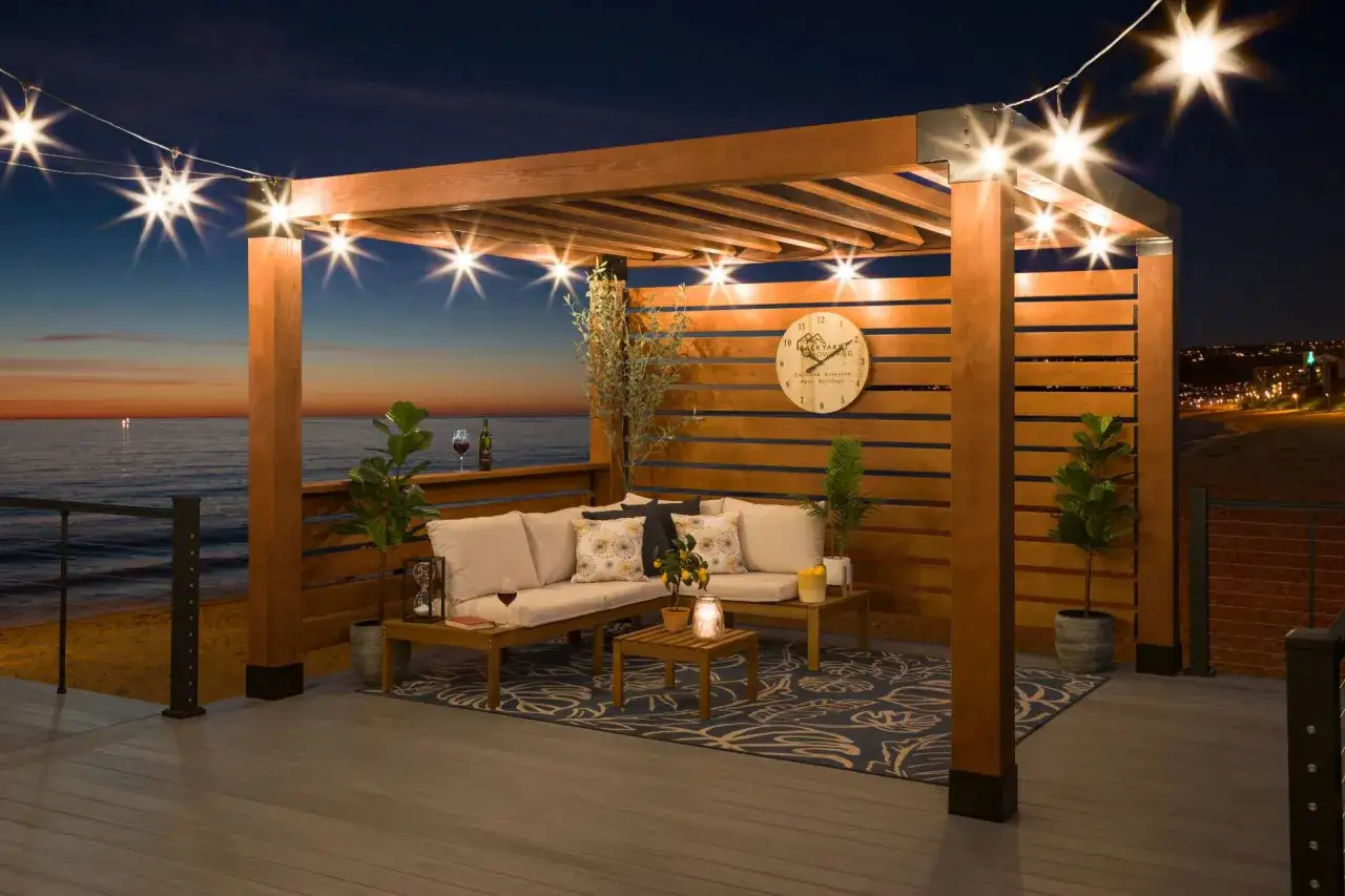 Ambiance nocturne sur une terrasse avec une pergola en bois. Un salon ext&eacute;rieur confortable invite &agrave; la d&eacute;tente, avec vue sur la mer et les lumi&egrave;res de la ville. Id&eacute;al pour construire une pergola et profiter de ces moments.