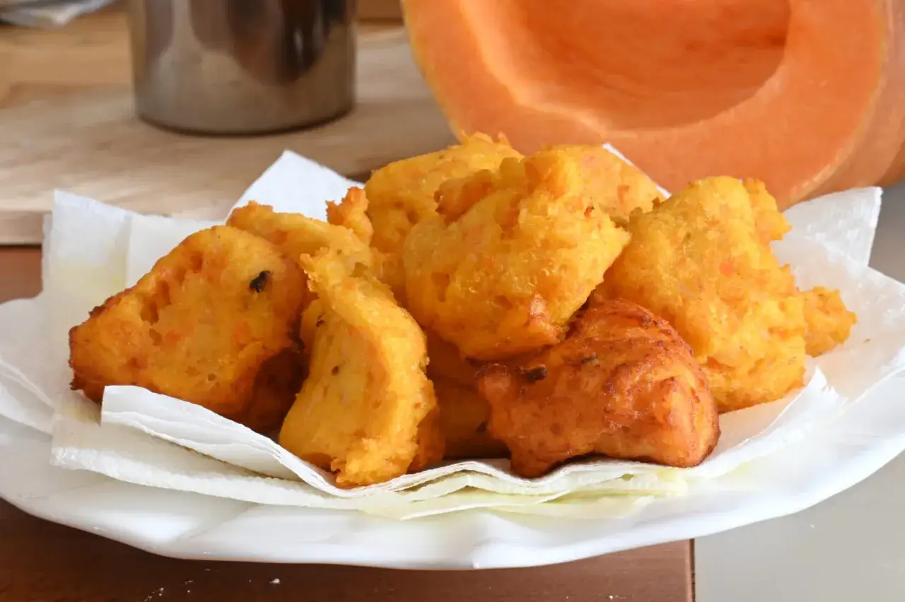 Frittelle di zucca dorate e croccanti, servite su un piatto bianco con tovaglioli di carta. Sullo sfondo, una fetta di zucca arancione.
