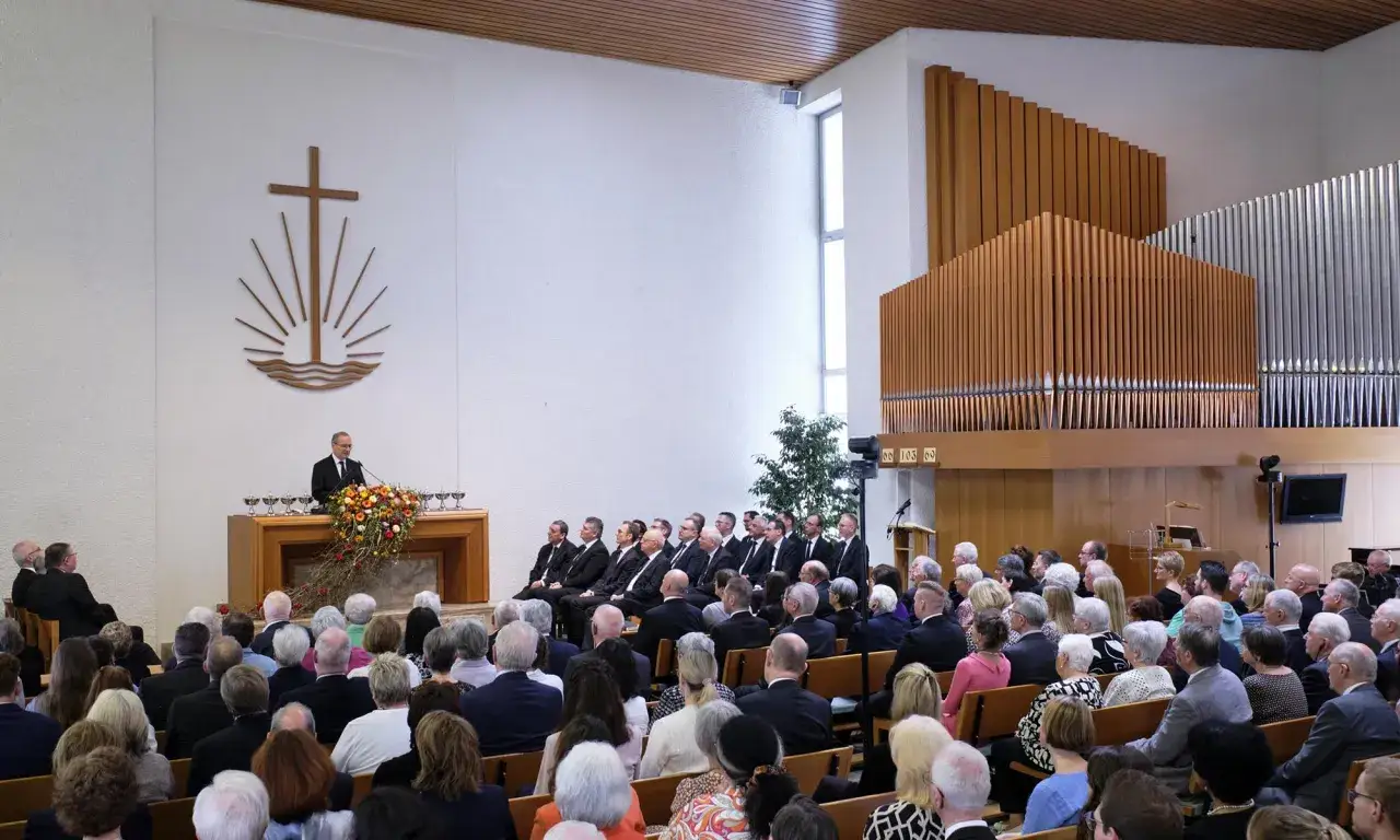 Predigt in der neuapostolischen Kirche. Prominente Mitglieder sitzen im Publikum, während ein Geistlicher am Pult spricht.