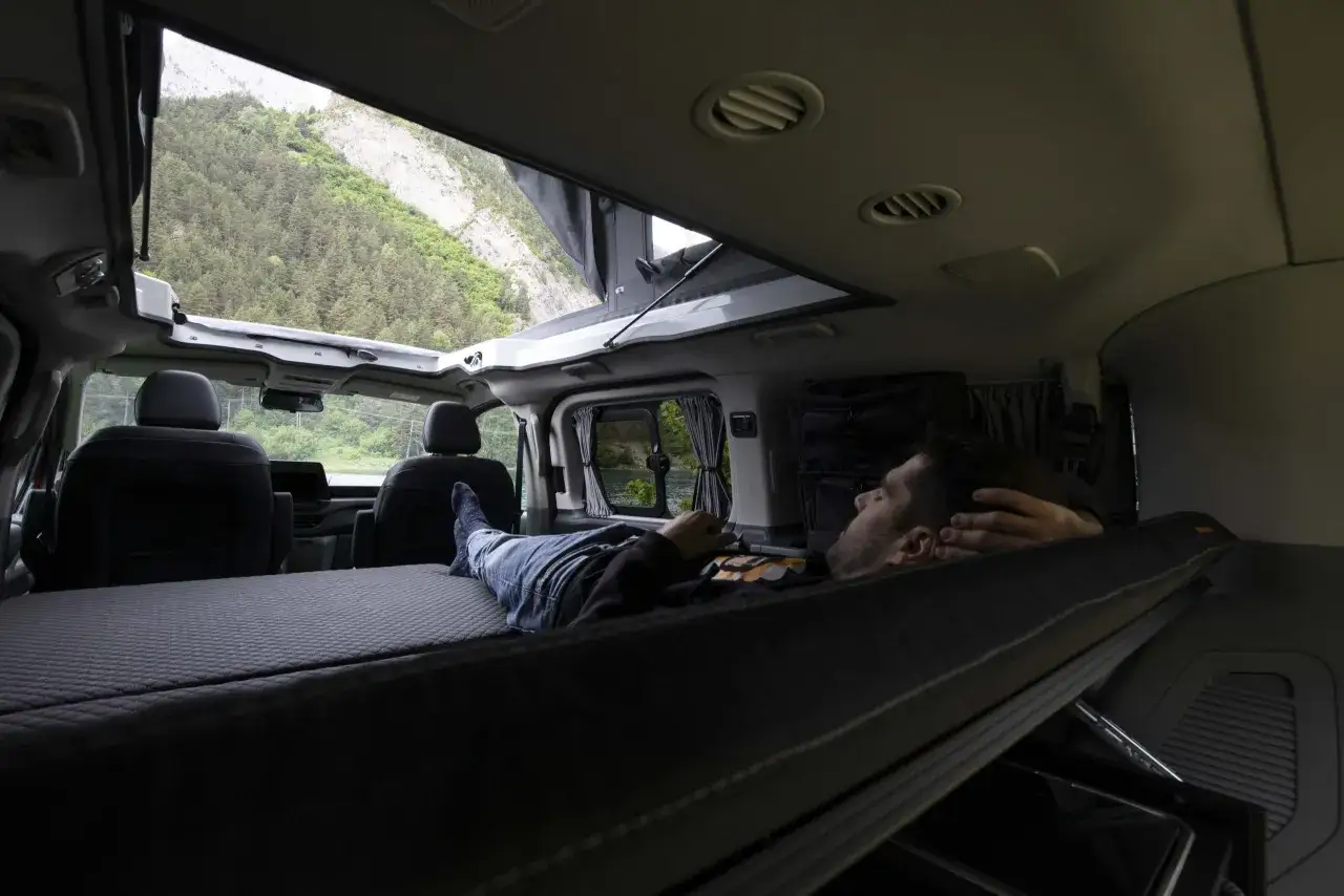 Hombre durmiendo en una cama improvisada en una furgoneta sin camperizar, con vistas a monta&ntilde;as y bosque.