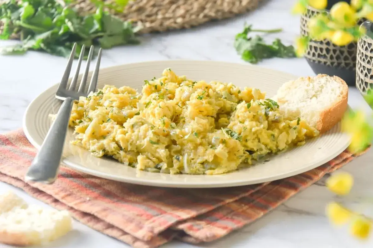 Delicioso revuelto de bacalao con perejil fresco, servido con una rebanada de pan r&uacute;stico y un tenedor listo para disfrutar.