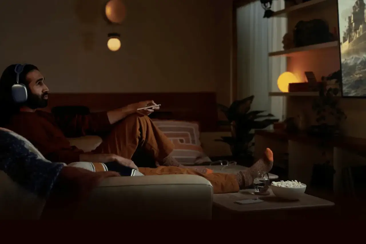 Mężczyzna w słuchawkach z technologią airplay relaksuje się na kanapie, oglądając film na telewizorze. Obok miska popcornu.