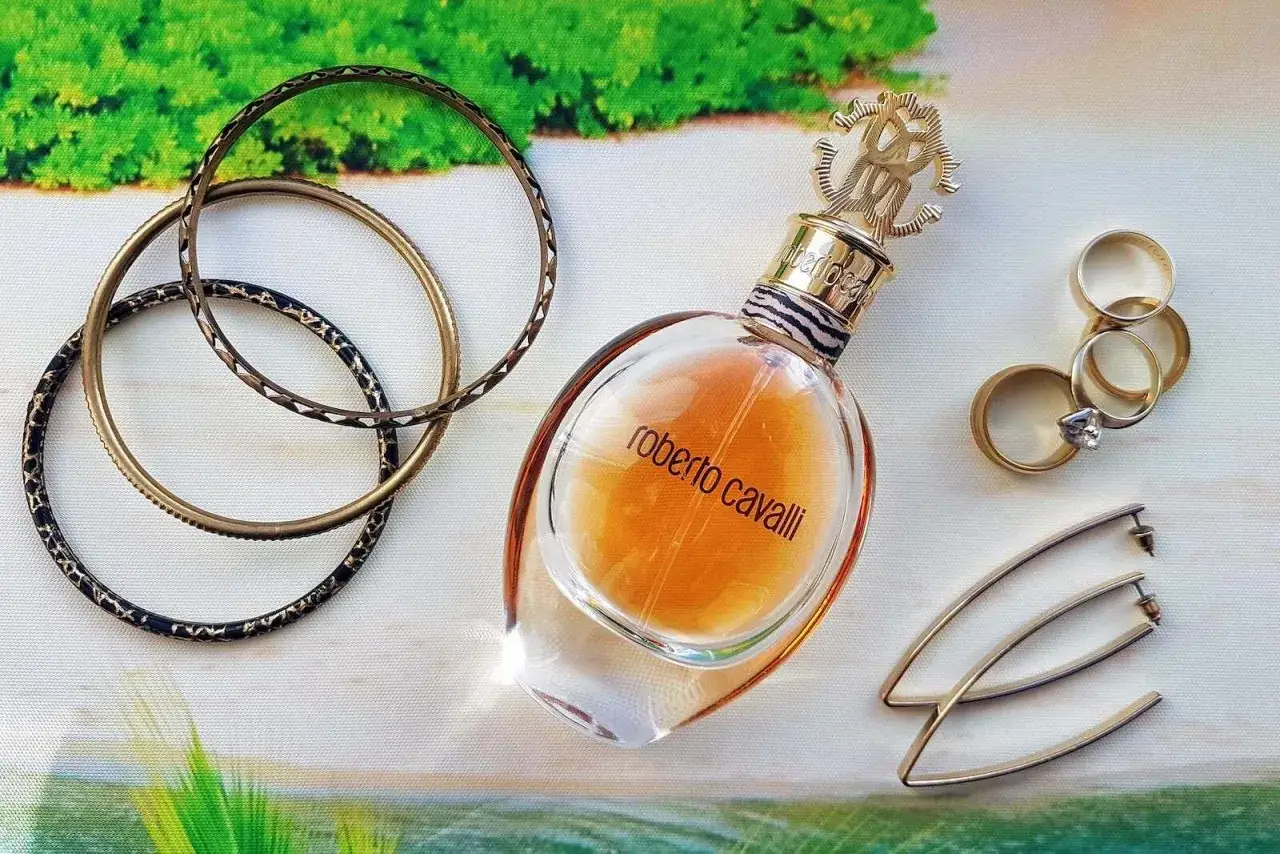 Złota butelka roberto cavalli edp, bransoletki i pierścionki na tle tropikalnej plaży.