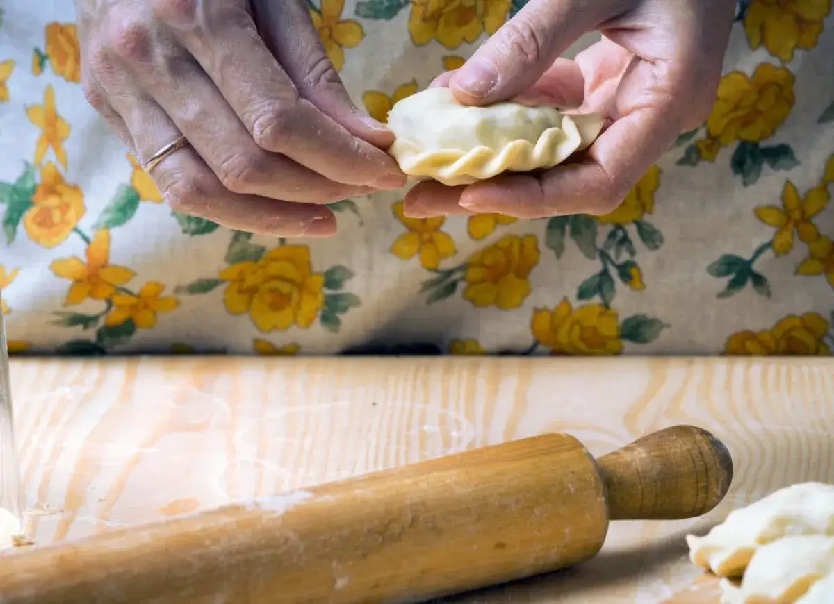 Ręce formują pieroga, przygotowując go do gotowania. W tle fartuch z żółtymi kwiatami i wałek do ciasta. Idealny przepis na ciasto na pierogi.