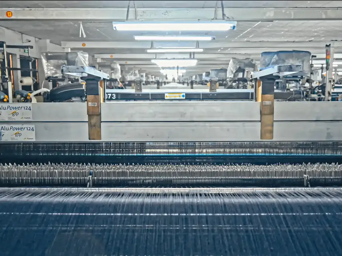 Maszyna tkacka tworzy tkaninę, z której powstaje jeans. Widać rzędy nici i mechanizmy maszyny.