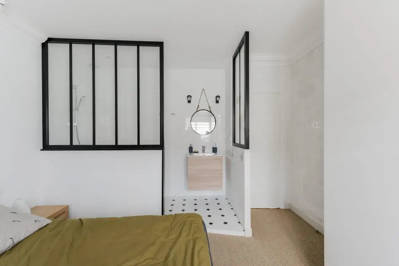 Chambre parentale avec salle de bain ouverte. Douche vitr&eacute;e, lavabo et miroir rond. Lit avec couette kaki.