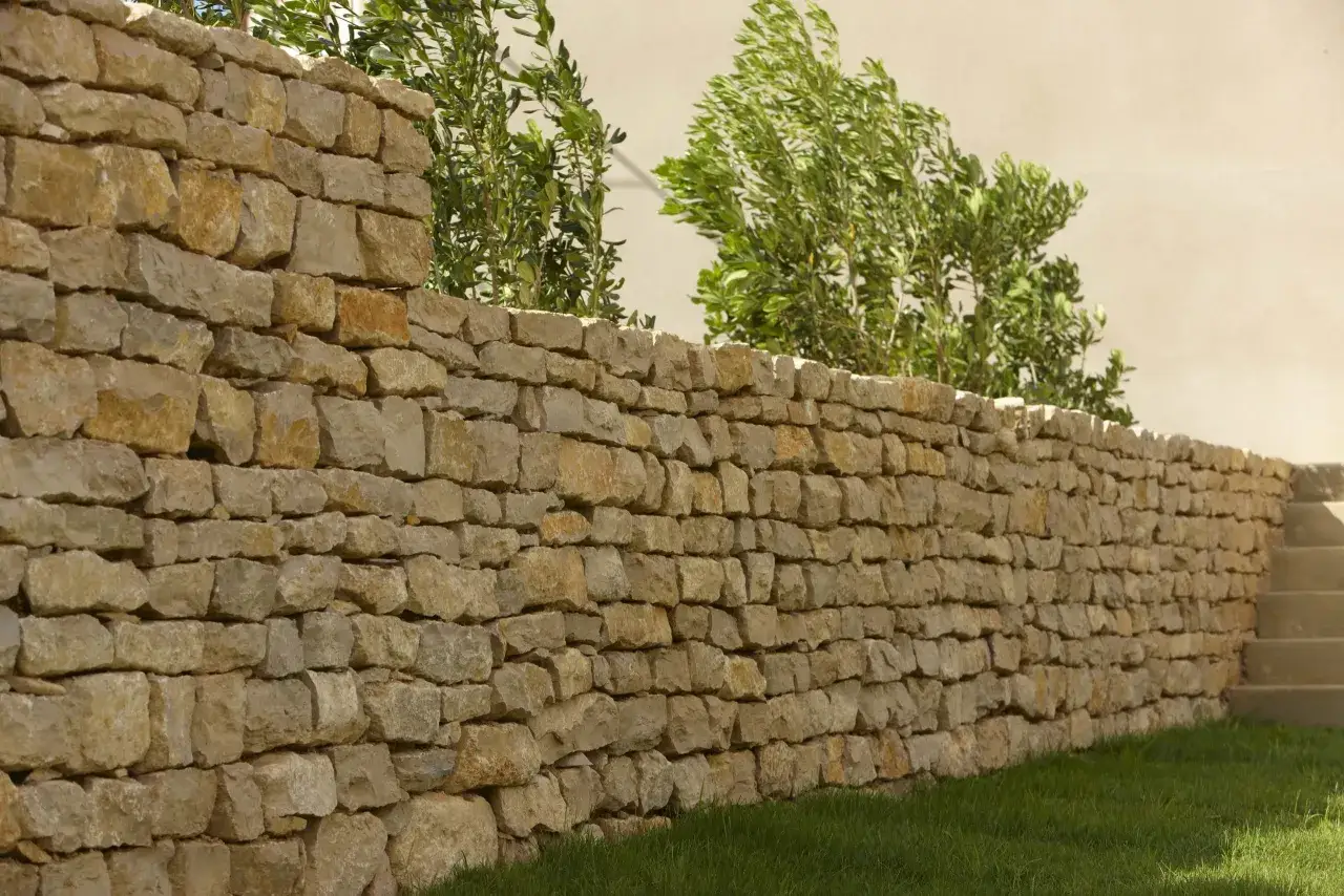 Un mur en pierre sèche, bordé d'herbe verte et d'arbustes luxuriants, s'étend sur la droite, menant à des marches en pierre.