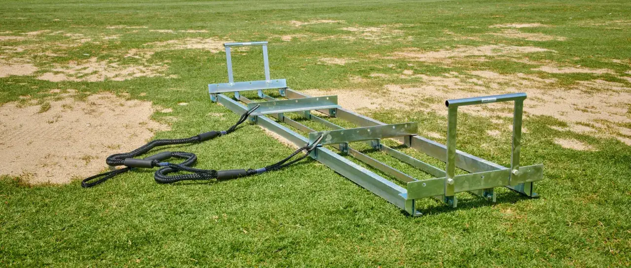 Metalowy aerator z linami, idealny do tego, jak wyrównać trawnik. Narzędzie leży na zielonej trawie i piasku.