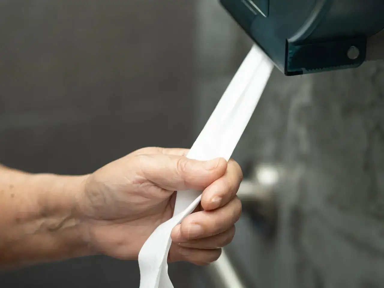 Dłoń zrywa papier z dozownika. Uchwyt na papier toaletowy zamontowany jest na wygodnej wysokości, dostępnej dla każdego.
