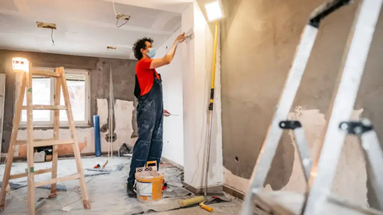 Un trabajador con mascarilla pinta una pared en una obra. Escaleras y cubos de pintura sugieren que se está aplicando el **artículo 22 de la L.A.U.**