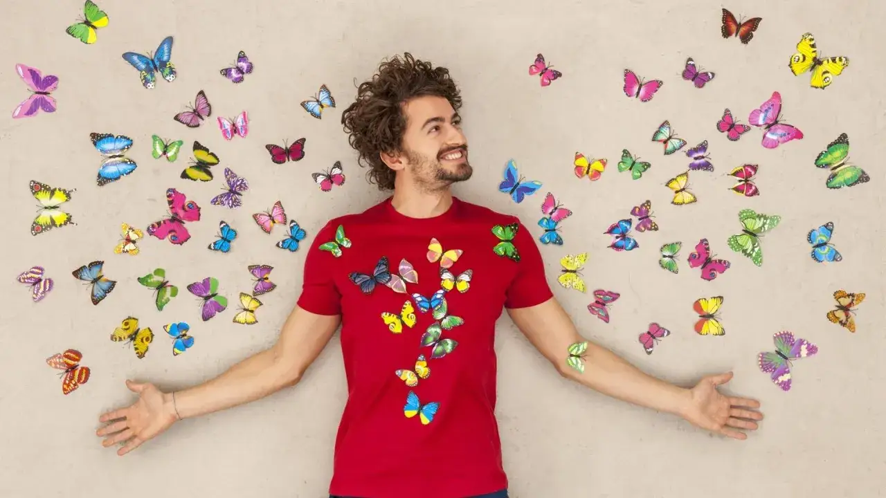 Ein Mann in einem roten T-Shirt, umgeben von bunten Schmetterlingen, die ihn wie eine Vision umfliegen. Er blickt nach oben, als w&uuml;rde er sich fragen: warum lebe ich?