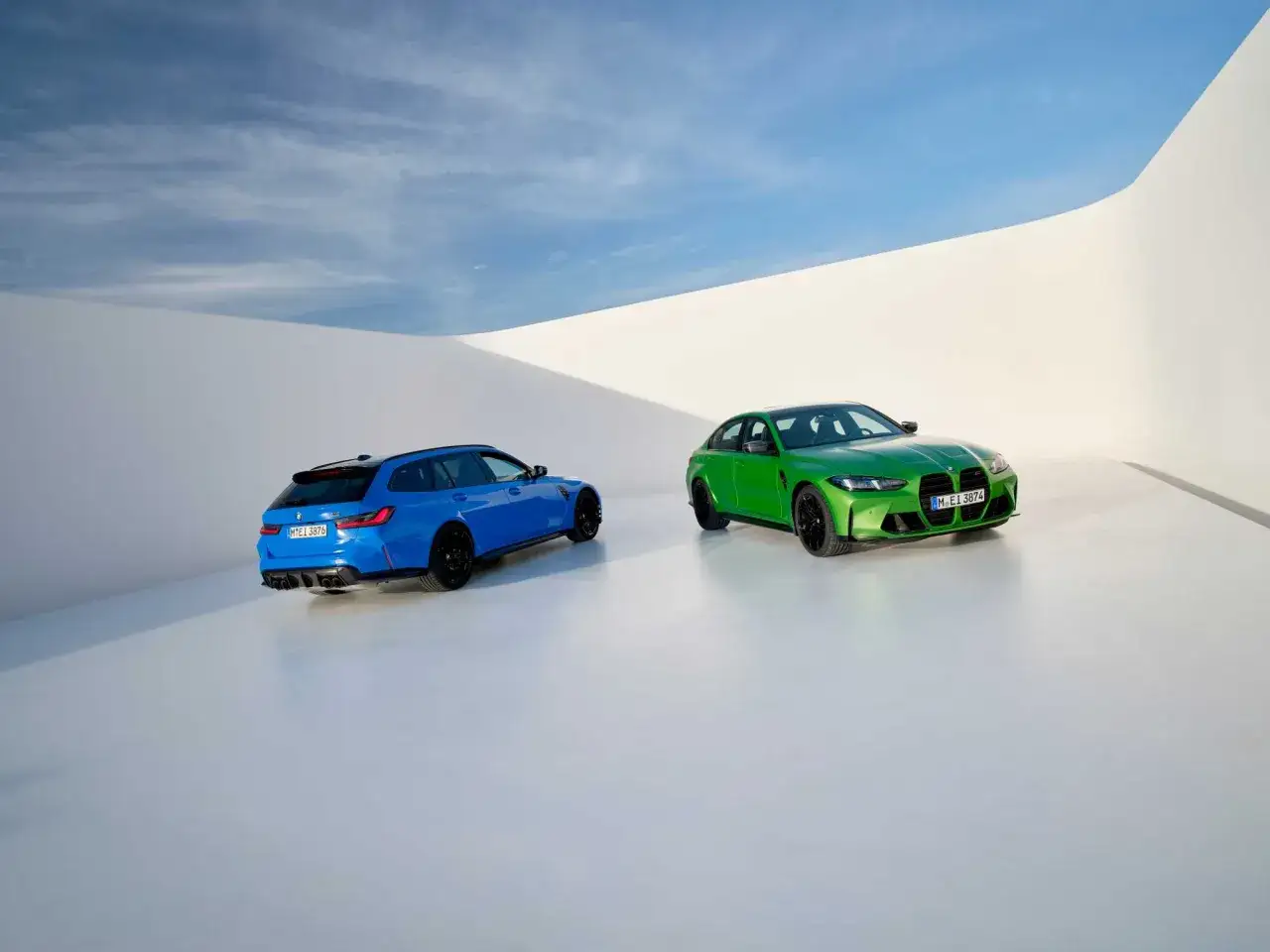 Dwa sportowe BMW M 3: jeden w kolorze niebieskim kombi, drugi zielony sedan, stoją na białej, minimalistycznej scenie pod błękitnym niebem.