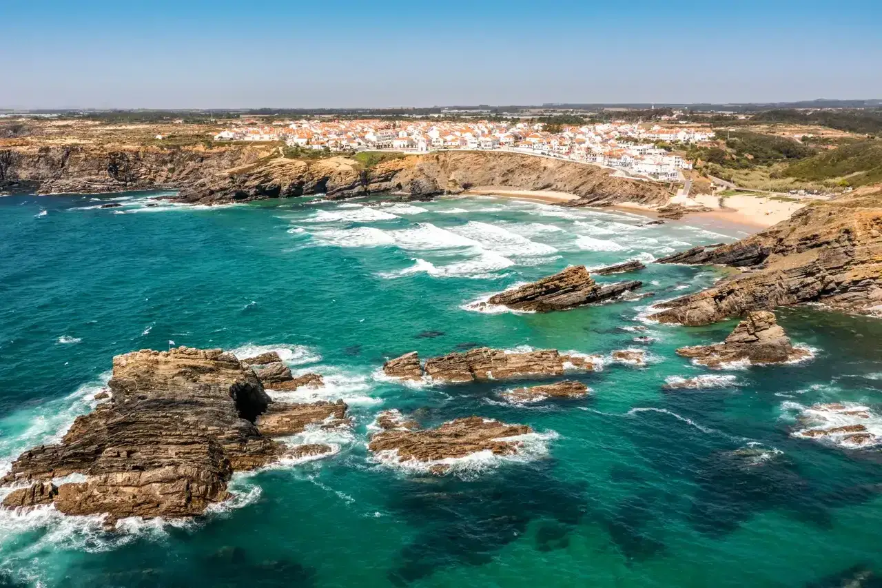 Costa rocosa y aguas turquesas en una **playa Portugal**. Un pueblo blanco se asoma al acantilado.