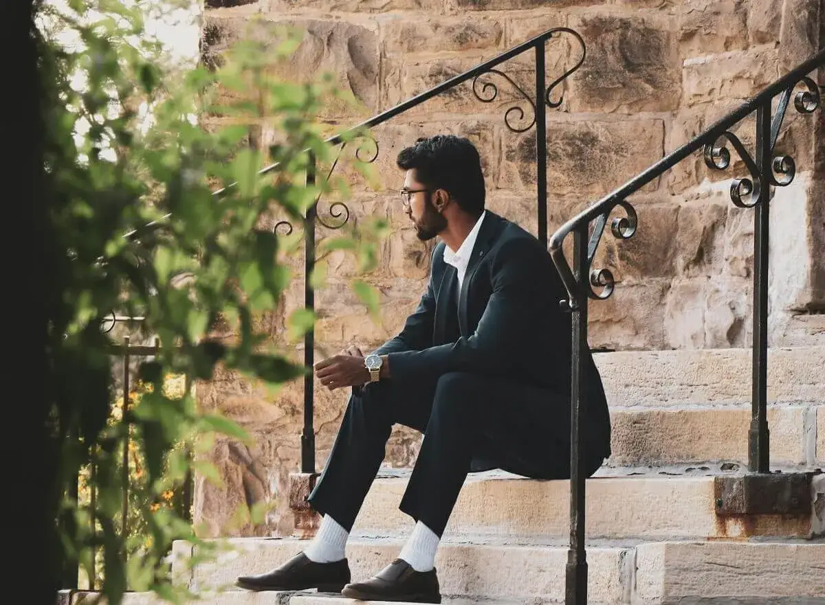 A man in a black suit and brown shoes sits on stone steps, looking thoughtfully into the distance.