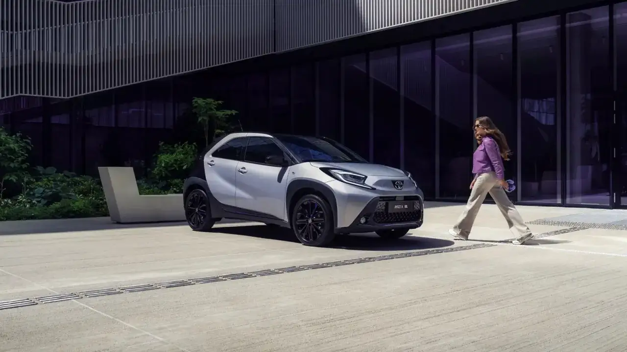 Elegancka kobieta mija srebrne Toyota Aygo X. Kompaktowe wymiary tego auta sprawiają, że idealnie wpasowuje się w miejski krajobraz.