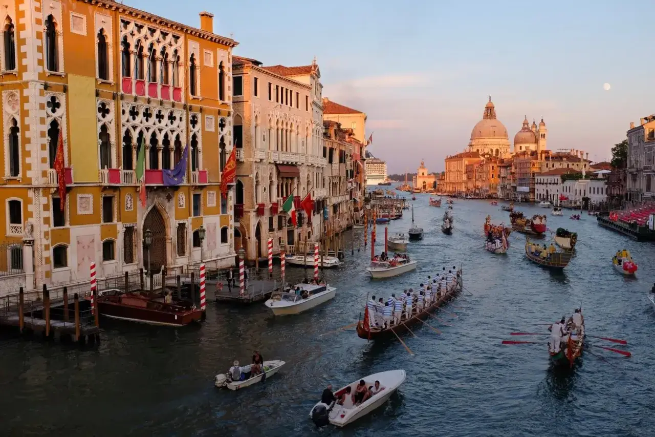 La Regata Storica anime le eaux v&eacute;nitiennes. Des gondoles et des bateaux traditionnels naviguent sur le Grand Canal, sous le regard des palais orn&eacute;s de drapeaux.
