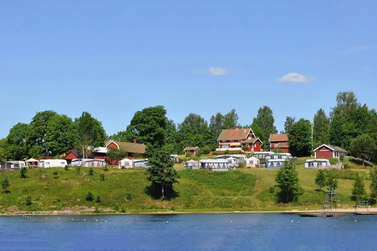 Gr&ouml;na kullar med husvagnar och r&ouml;da stugor vid en sj&ouml;, en idyllisk plats f&ouml;r greans camping.