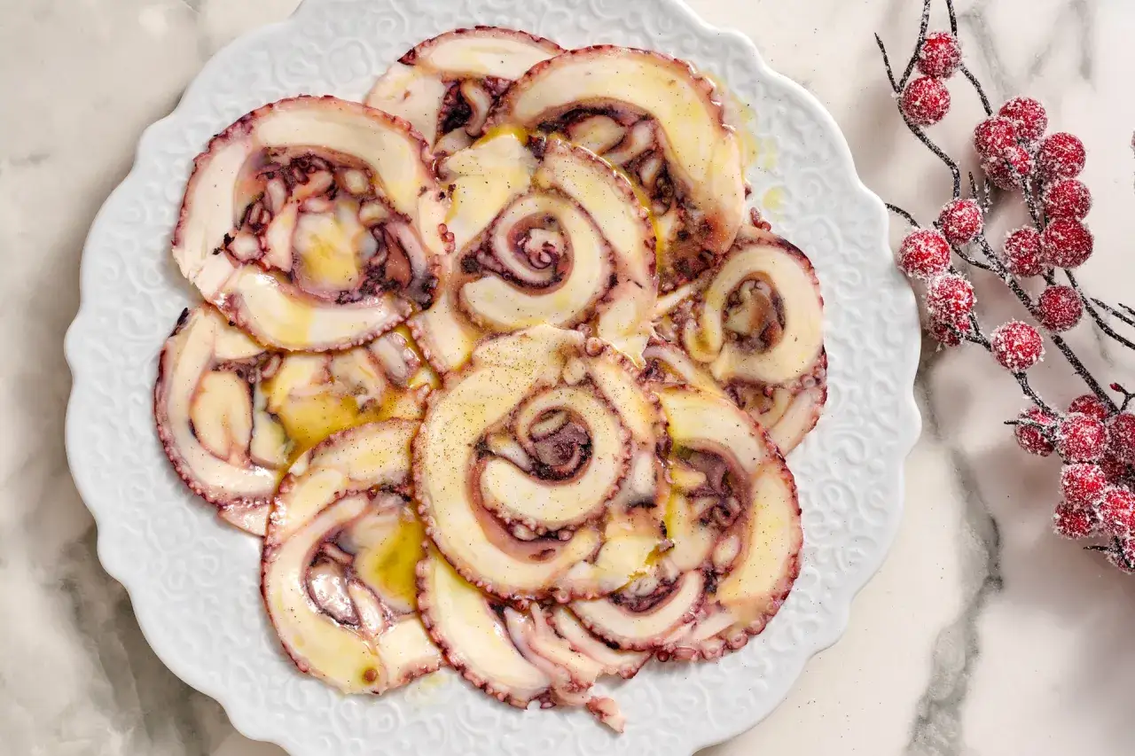 Fette di carpaccio di polpo condite con olio e pepe, disposte su un piatto bianco decorato. Accanto, un ramo con bacche rosse innevate.